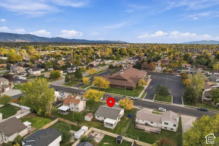 Aerial perspective featuring a residential lot with a paved driveway, attached garage, covered rear patio, and mature trees, set against a mountainous horizon