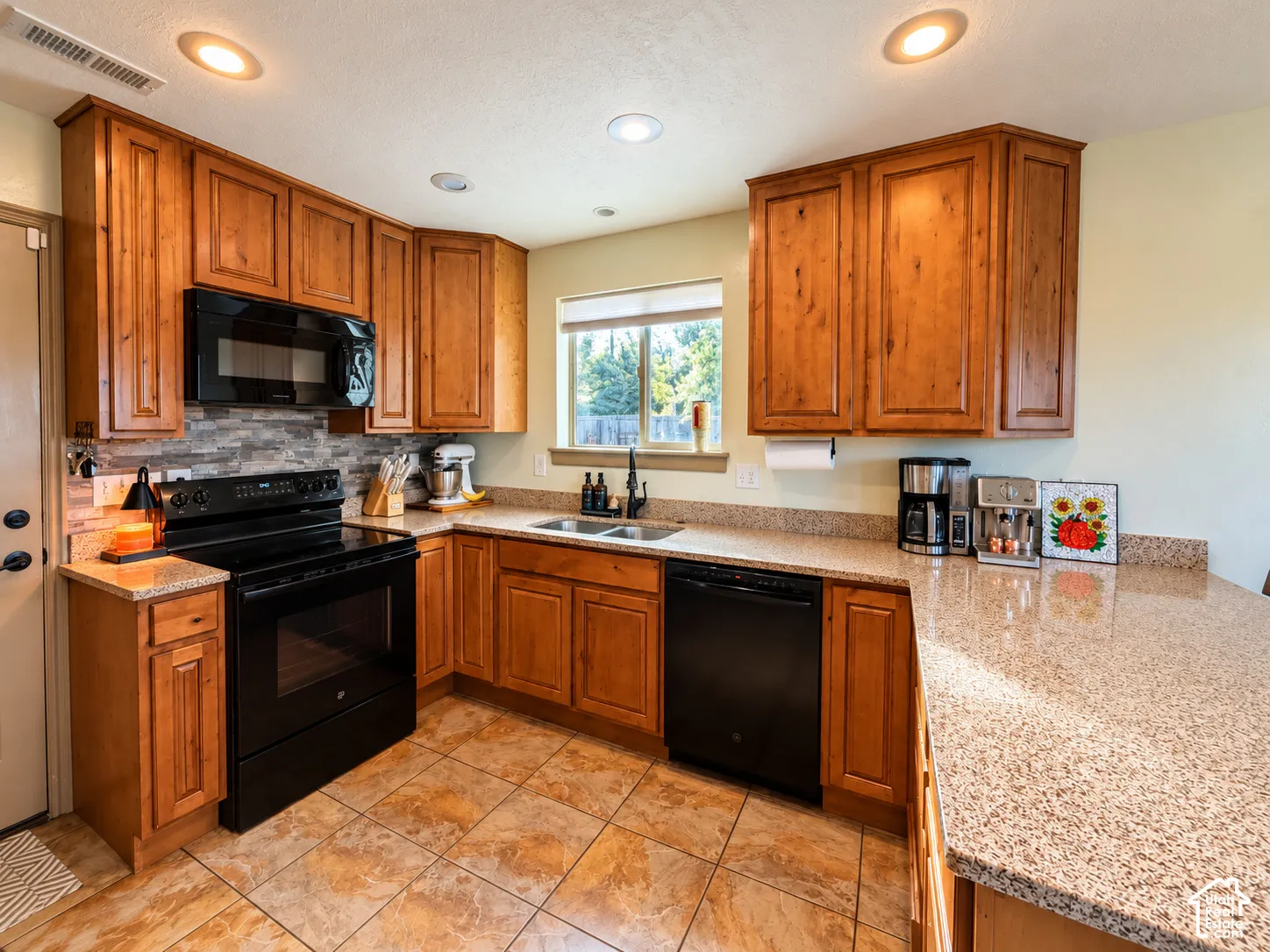 Kitchen featuring stained wood cabinetry, stone-style tile backsplash, solid-surface countertops, black appliances, and tile flooring