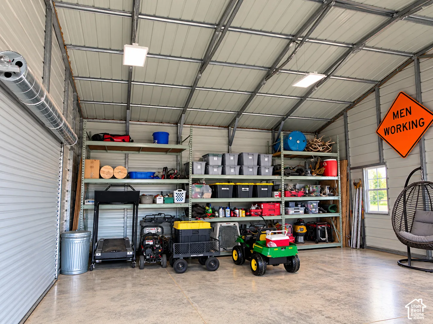 Detached workshop featuring a high-clearance metal roof, exposed steel structural framing, polished concrete flooring, integrated LED overhead lighting, and extensive wall-mounted industrial metal shelving