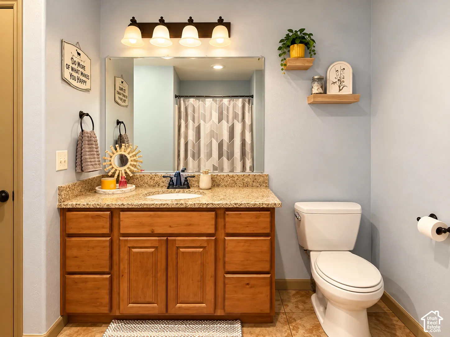 Wood-finish vanity cabinet, stone-finish countertop with an integrated oval sink, four-light vanity fixture, wall-mounted floating shelving, and tile flooring