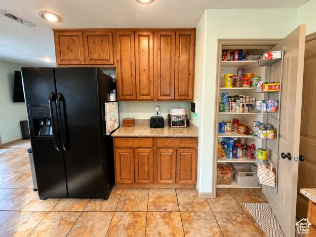 Kitchen featuring custom wood cabinetry, stone-finish countertops, black side-by-side refrigerator, tile flooring, and a built-in pantry with door-mounted storage racks
