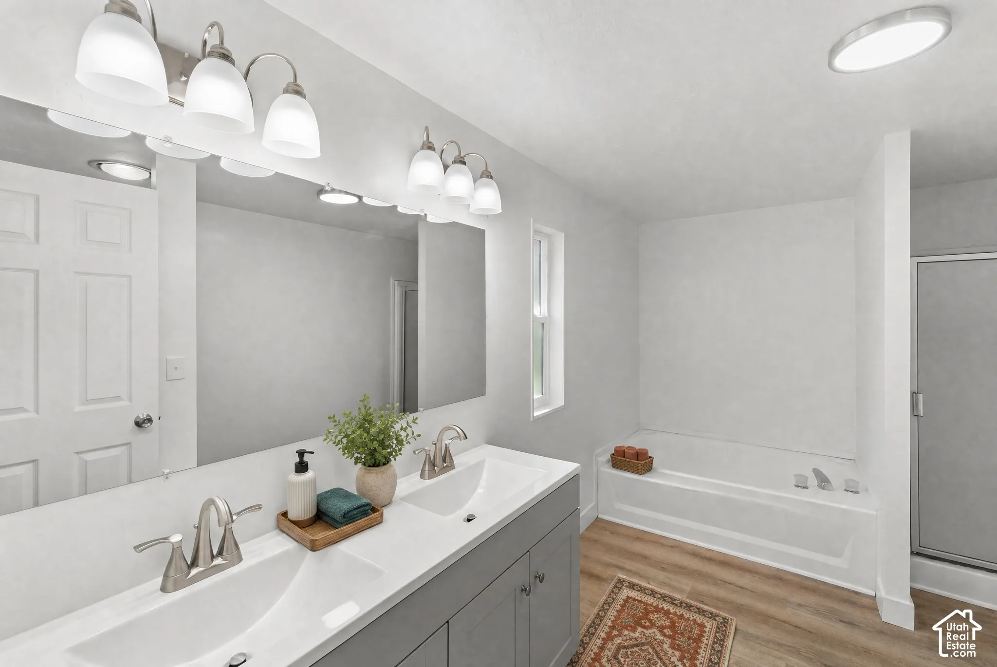 Dual-sink vanity with white countertops, expansive wall mirror, brushed metal hardware, soaking tub, and enclosed glass shower