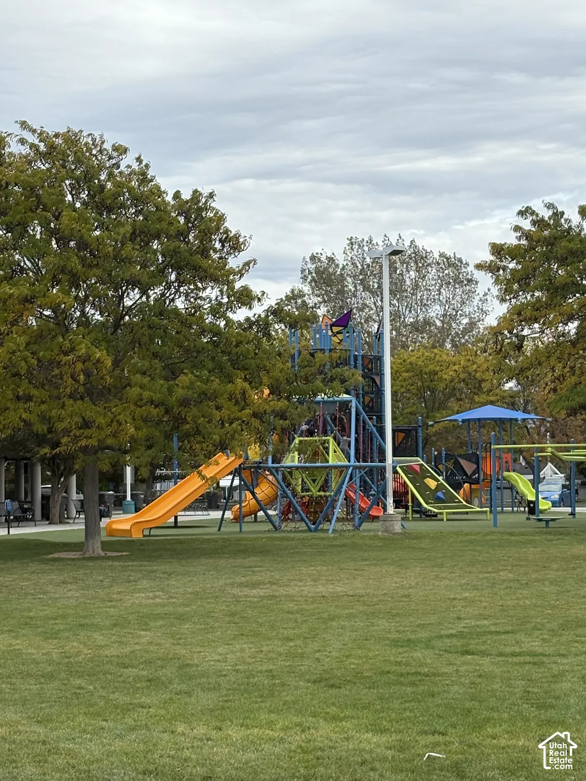 Outdoor recreational space featuring a multi-level playground structure, integrated slides, a climbing frame, and mature trees situated on a level grass area