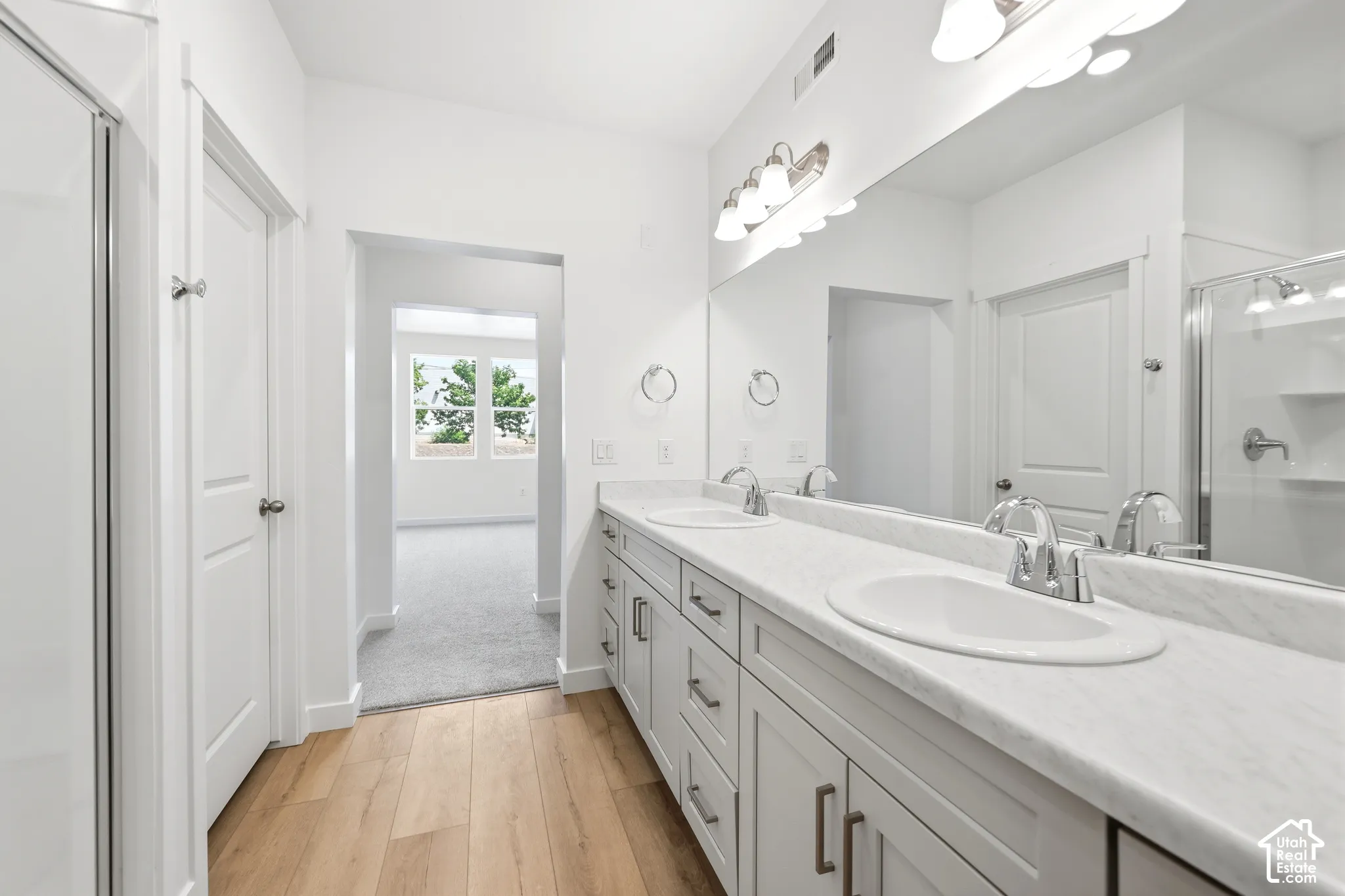 Dual-sink vanity with white stone-finish countertop, cabinetry, and full-width vanity mirror, featuring wood-finish flooring and a glass-enclosed shower