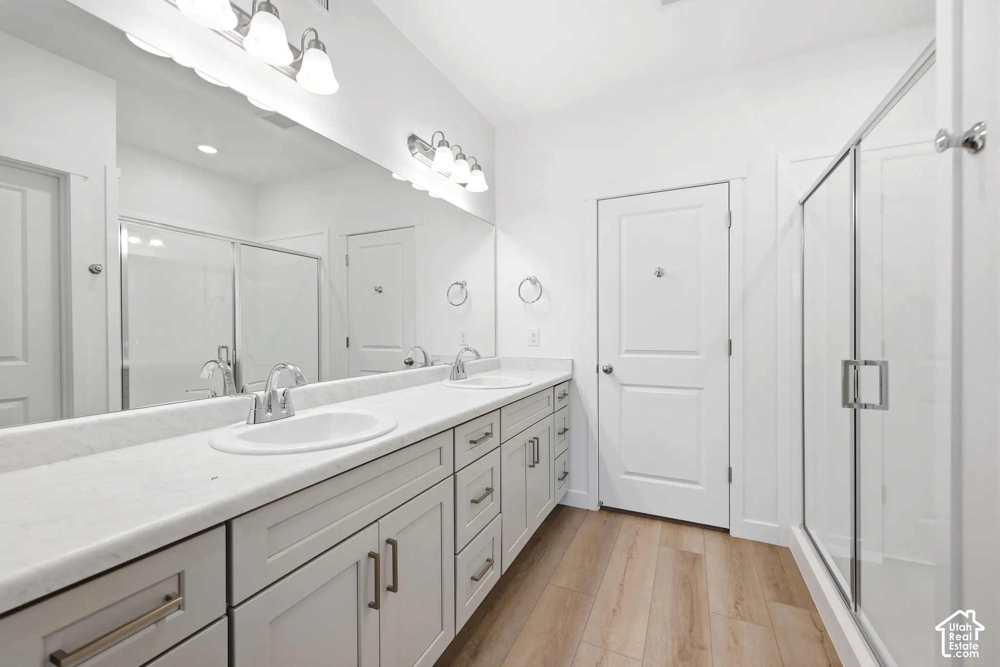 Dual-vanity cabinetry with light-toned countertops, full-width vanity mirror, walk-in shower with glass enclosure, and wood-finish flooring