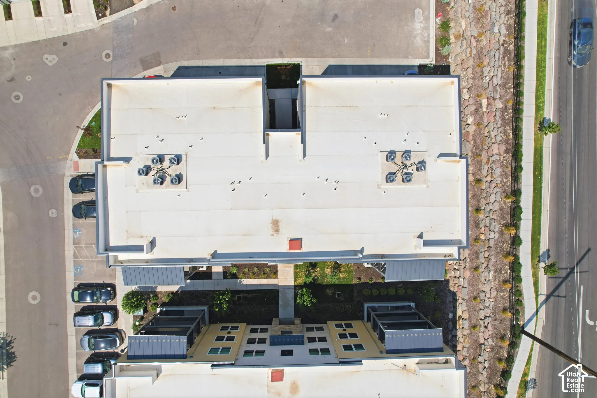 Aerial view of a multi-level structure featuring a flat roof system, rooftop HVAC units, stone retaining wall, paved surface parking, and manicured landscaping