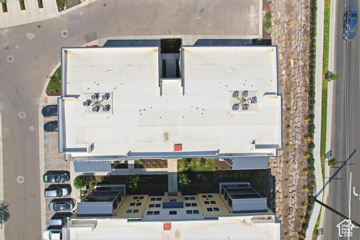 Aerial view of a multi-level structure featuring a flat roof system, rooftop HVAC units, stone retaining wall, paved surface parking, and manicured landscaping
