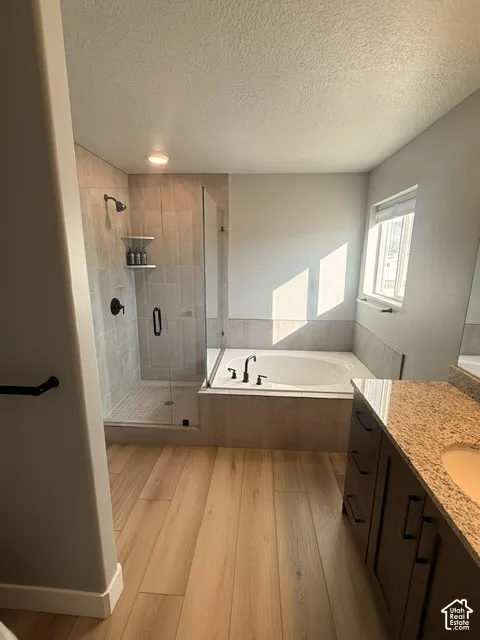 Bathroom featuring wood-finish flooring, a vanity with a light-toned stone countertop, an inset soaking tub with tiled surround, and a walk-in shower with a glass enclosure and matte black fixtures