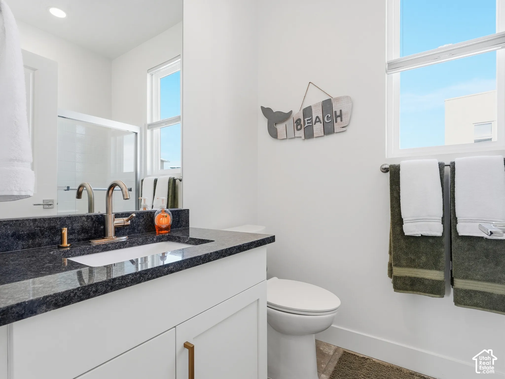 White cabinetry vanity with a dark stone countertop, undermount sink, metallic faucet, and large-format rectangular windows