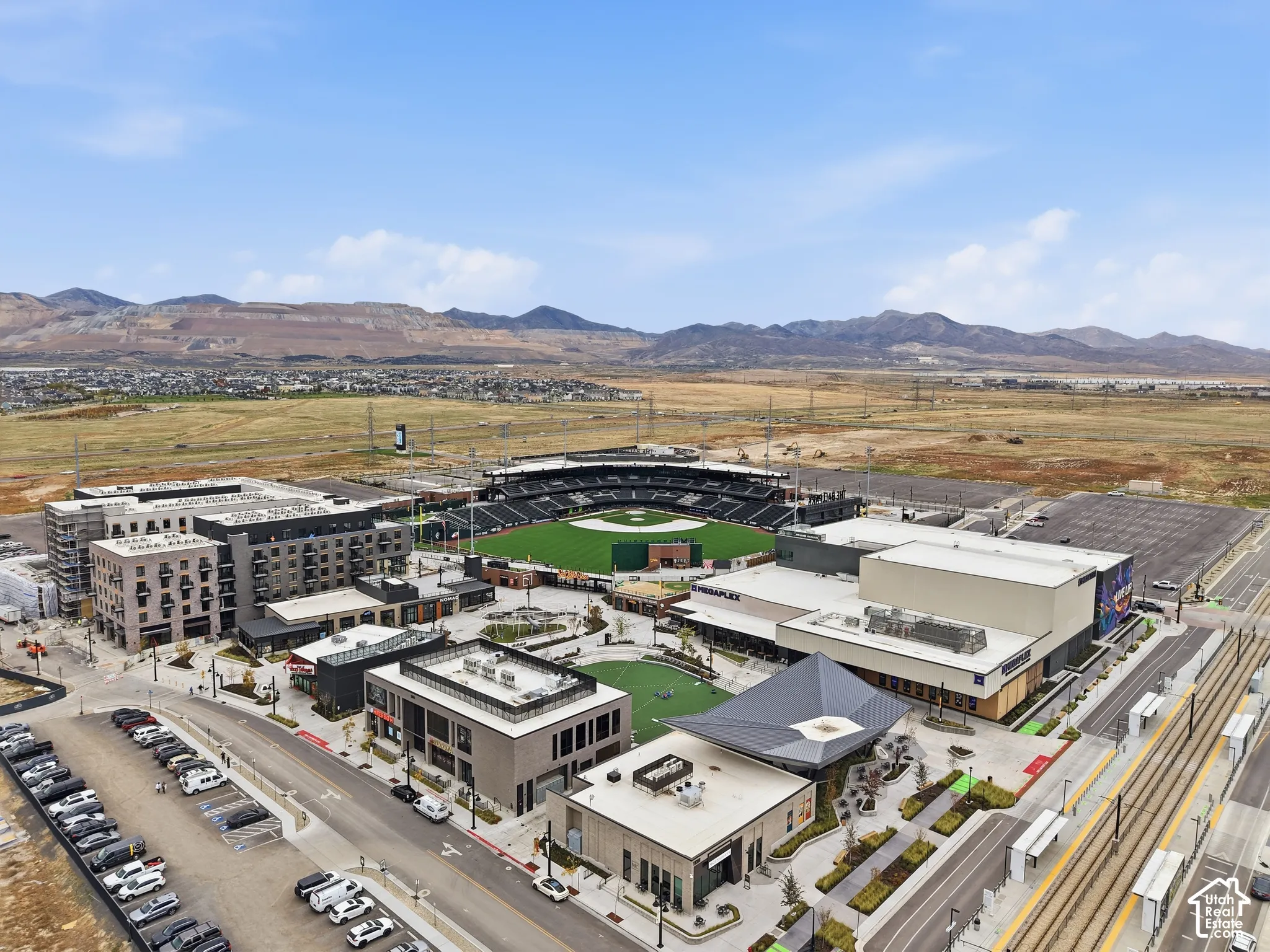 Aerial perspective of a multi-use complex featuring a professional stadium, structured parking, modern retail buildings, central green space, and proximity to light rail transit