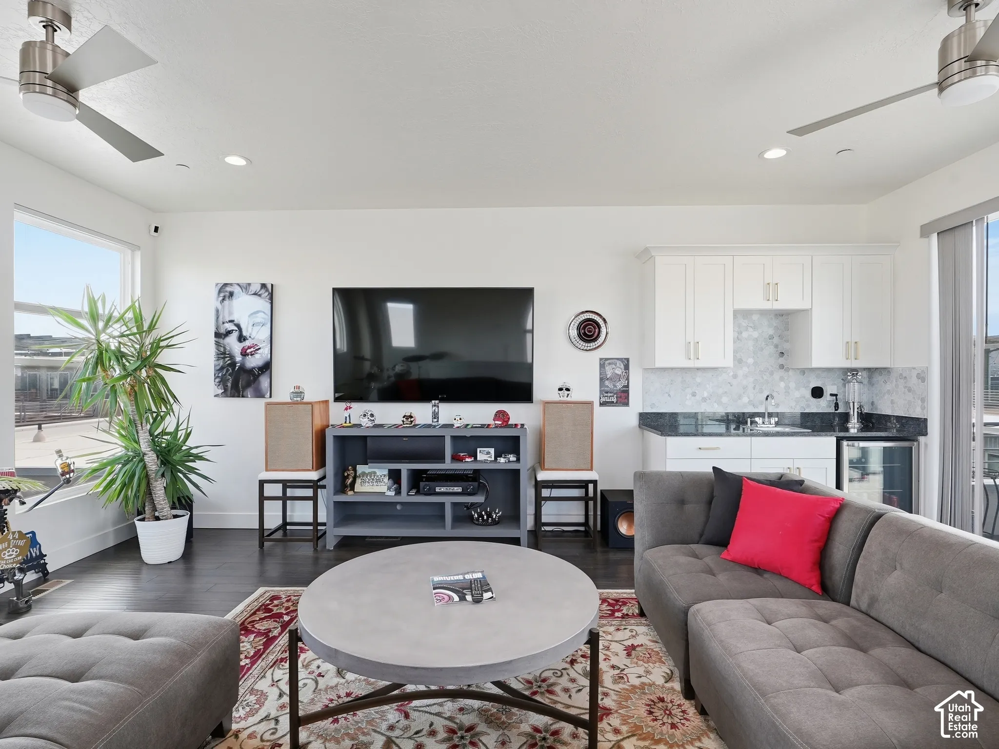 Living area featuring wood-finish flooring, dual brushed-nickel ceiling fans, recessed lighting, and a wet bar with white shaker-style cabinetry, stone-composite countertops, and a tiled backsplash