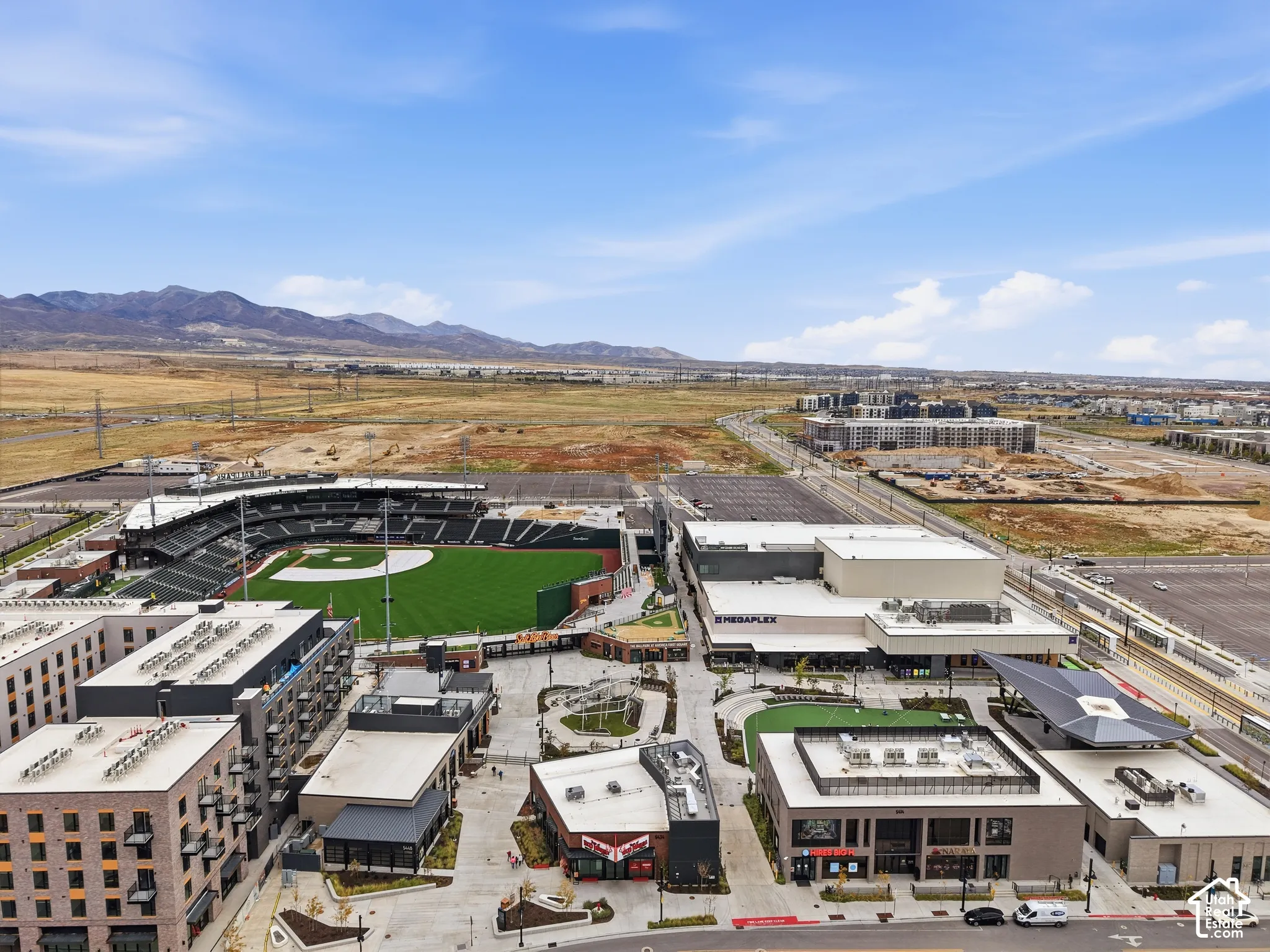 Aerial perspective of a multi-use complex featuring a professional baseball stadium, cinema, multi-story commercial buildings, paved pedestrian plazas, and a mountain range backdrop