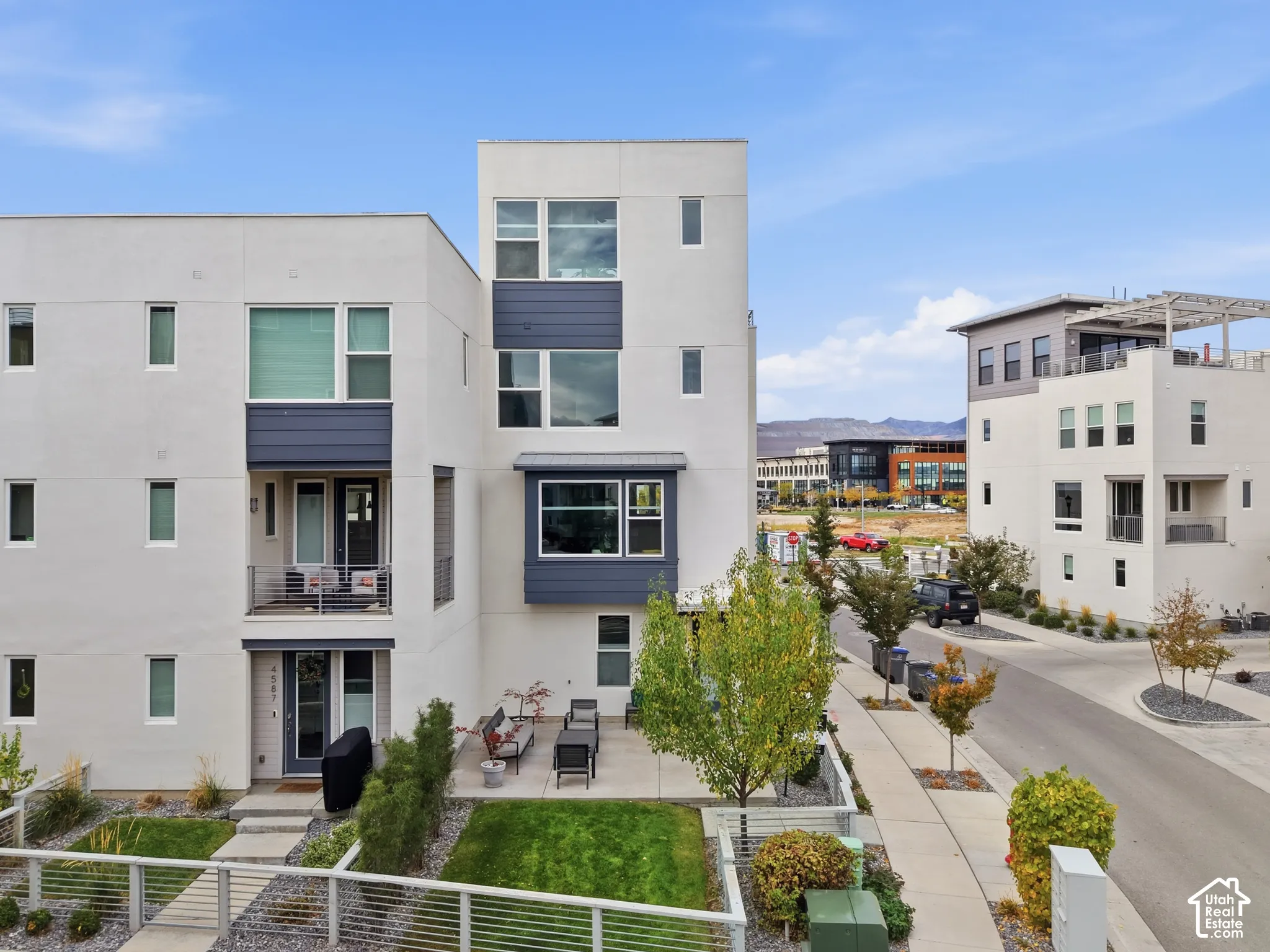 Modern multi-story exterior featuring light-toned smooth siding, dark horizontal accent panels, a covered ground-level patio, a second-story balcony with metal railings, and oversized rectangular windows
