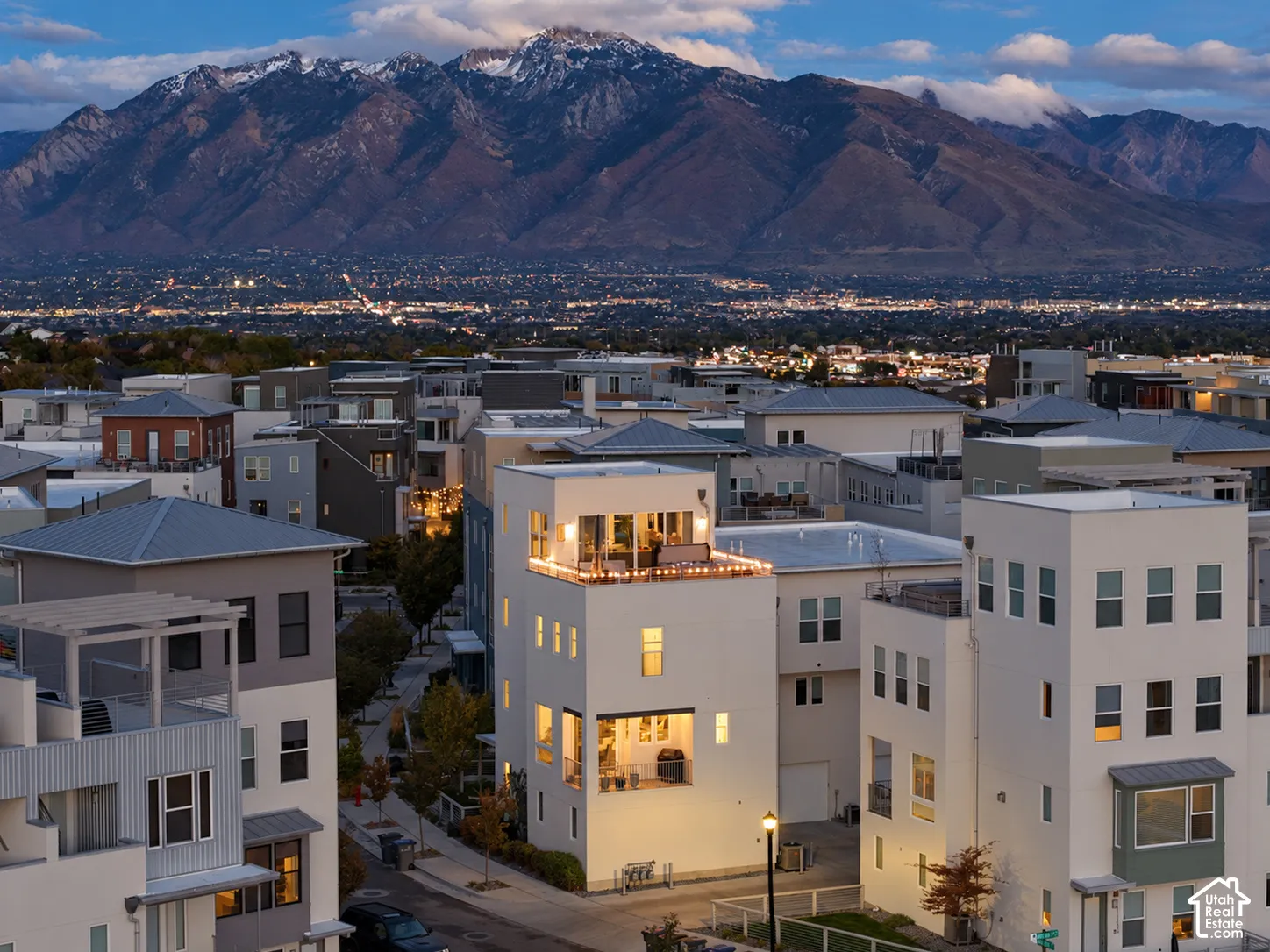 Modern multi-story structure featuring a rooftop deck and a recessed balcony, accented by a light-colored stucco exterior and a dramatic mountain backdrop