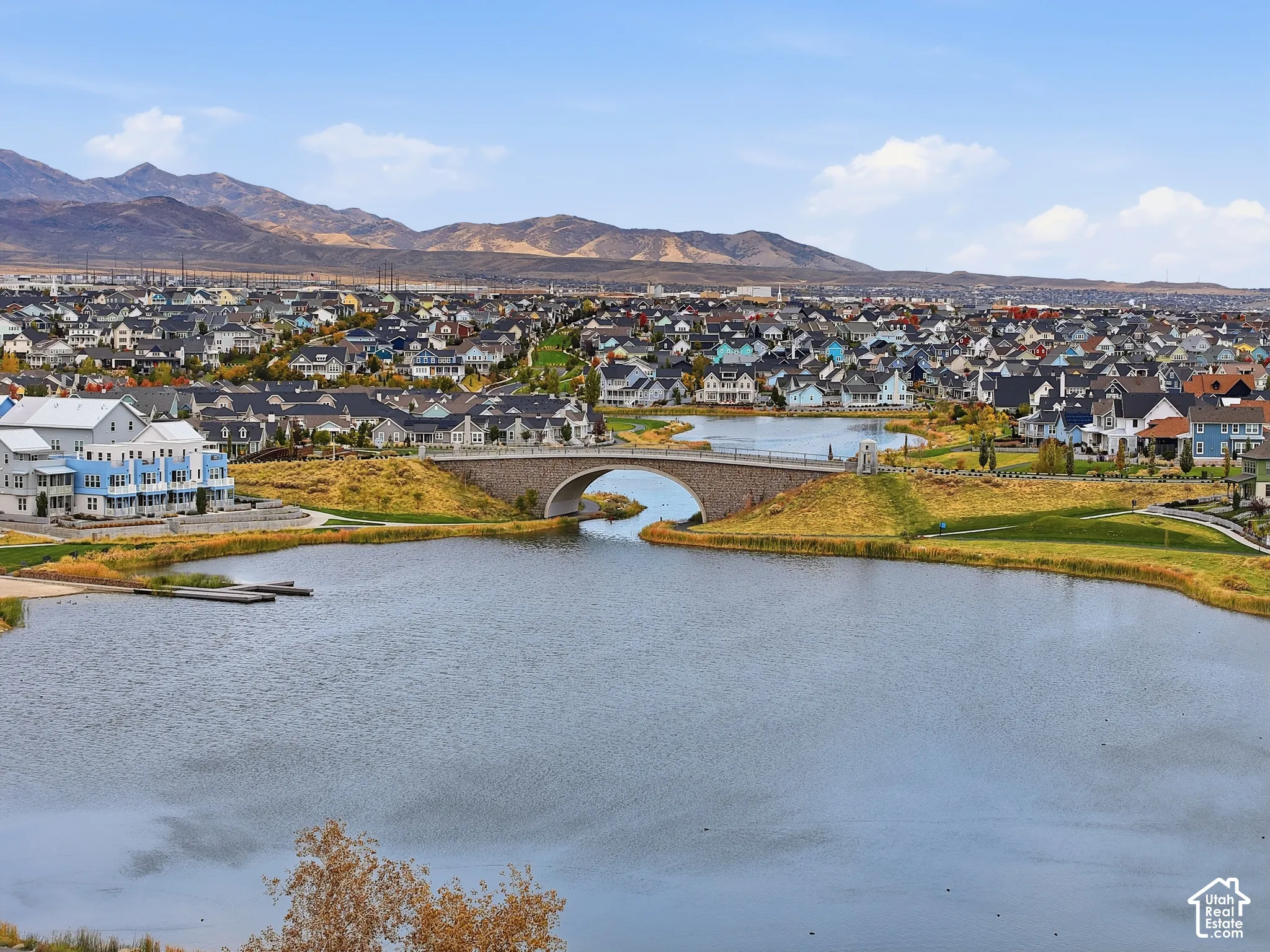 Aerial perspective of a lakefront community featuring a stone-arch bridge, expansive waterfront, paved walking paths, and mountain scenery