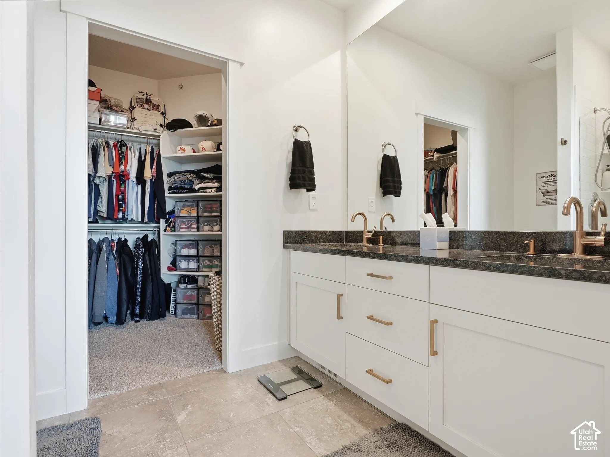 Dual-vanity cabinetry featuring white shaker-style doors, stone-finish countertops, and metallic hardware, paired with a full-width wall mirror and walk-in closet with built-in shelving