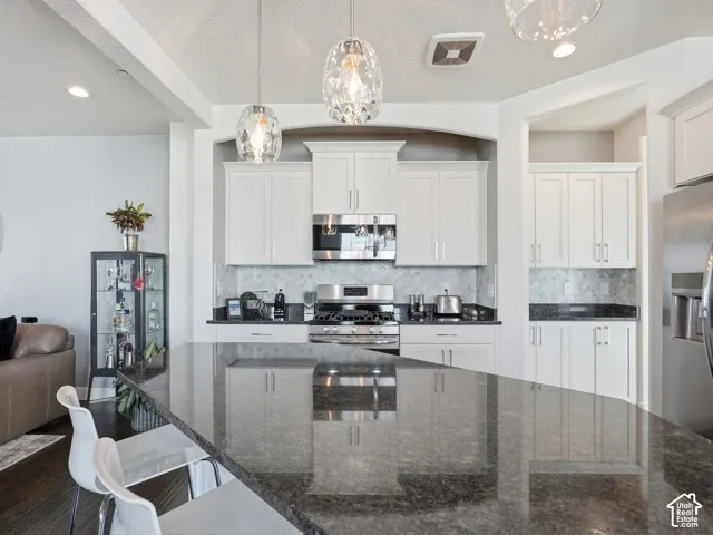 Kitchen featuring white cabinetry, dark stone countertops, stainless steel appliances, a tiled backsplash, and decorative pendant lighting