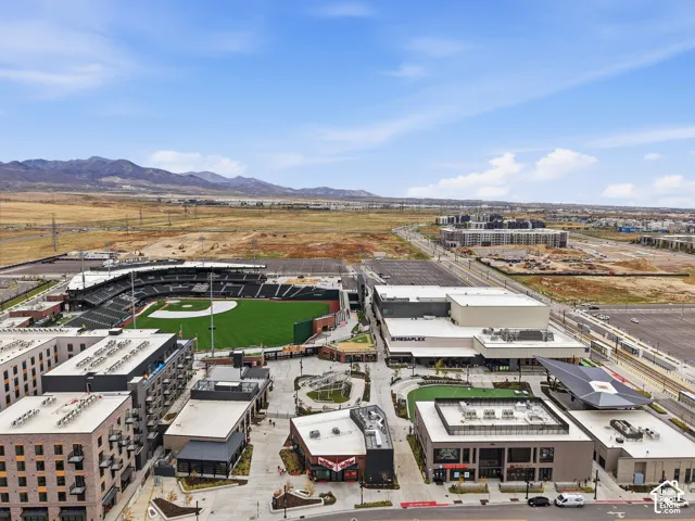 Aerial perspective of a multi-use complex featuring a professional baseball stadium, cinema, multi-story commercial buildings, paved pedestrian plazas, and a mountain range backdrop