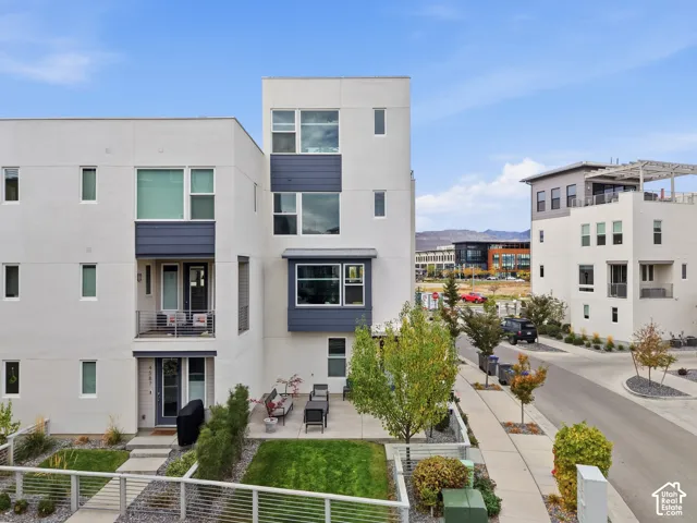 Modern multi-story exterior featuring light-toned smooth siding, dark horizontal accent panels, a covered ground-level patio, a second-story balcony with metal railings, and oversized rectangular windows