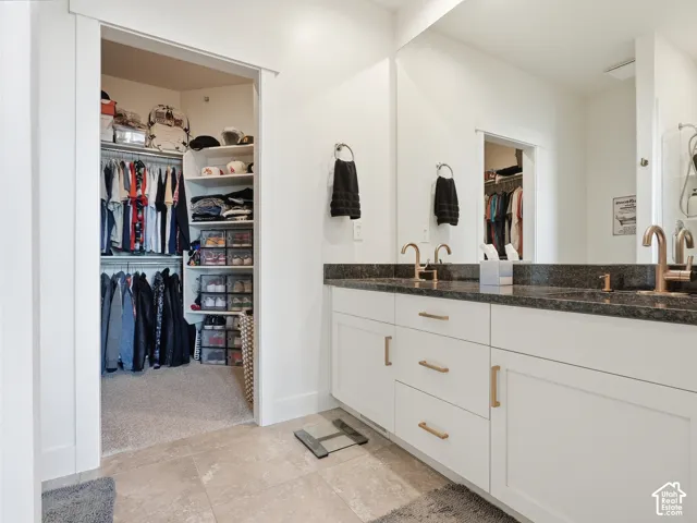 Dual-vanity cabinetry featuring white shaker-style doors, stone-finish countertops, and metallic hardware, paired with a full-width wall mirror and walk-in closet with built-in shelving