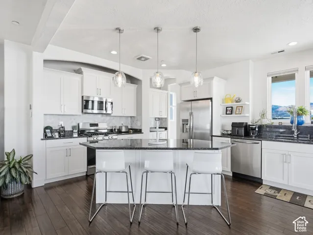 Kitchen featuring white cabinetry, dark stone countertops, stainless steel appliances, dark wood-finish flooring, and a central island with three glass pendant lights