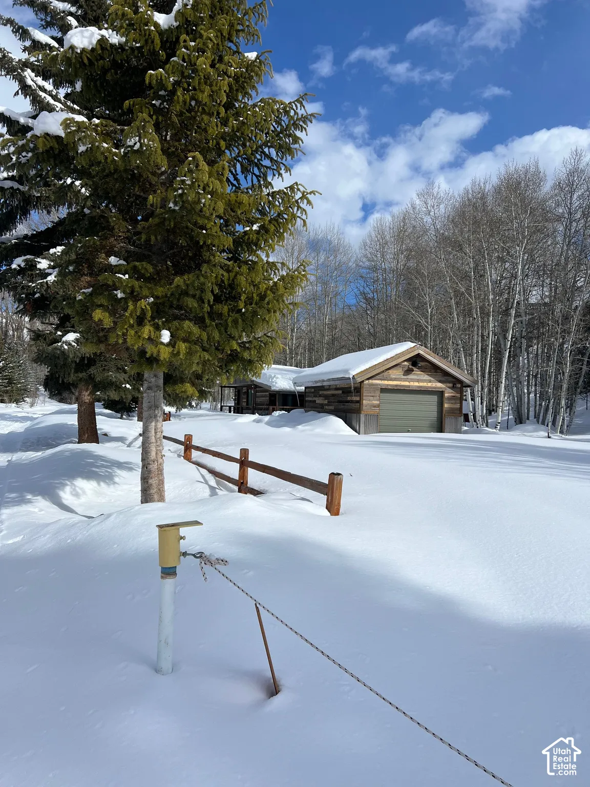 Wood-sided structure with a gabled roof and detached garage, featuring post-and-rail fencing amidst mature pine and aspen trees