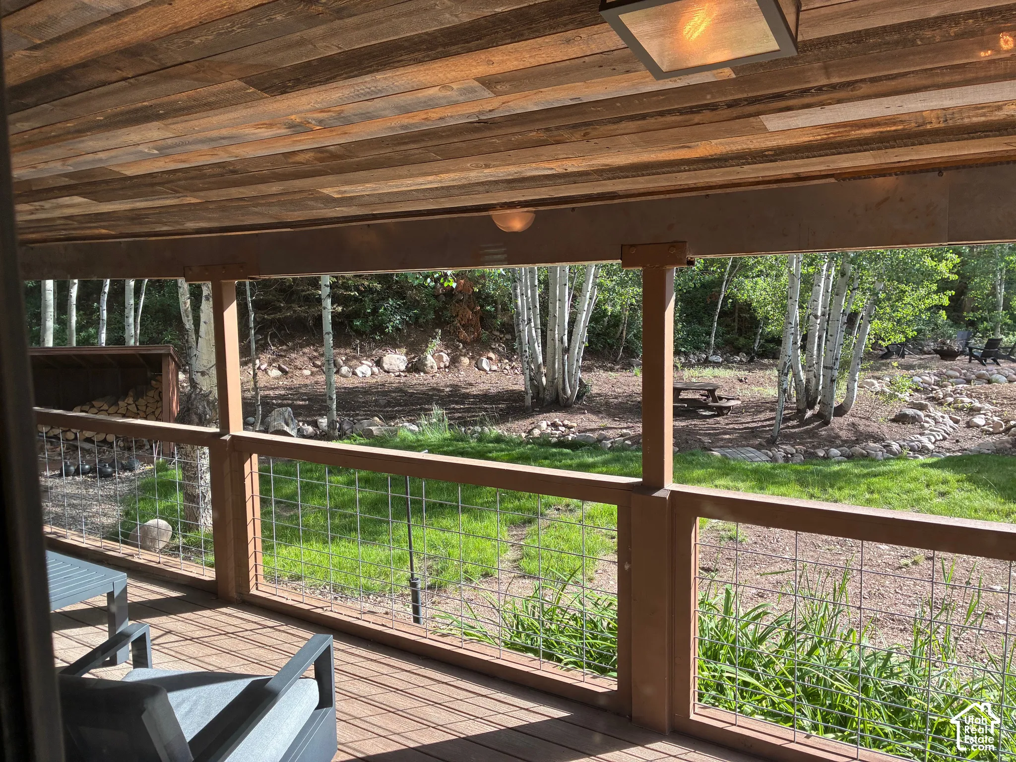 Covered deck featuring a natural wood-plank ceiling, wire-mesh railing, and exterior lighting, overlooking a landscaped yard with mature trees