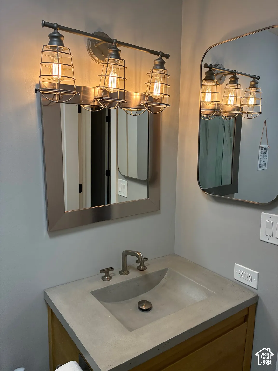 Industrial-style three-light vanity fixture with wire cages, rectangular mirror with metallic finish, integrated concrete basin, and wall-mounted faucet