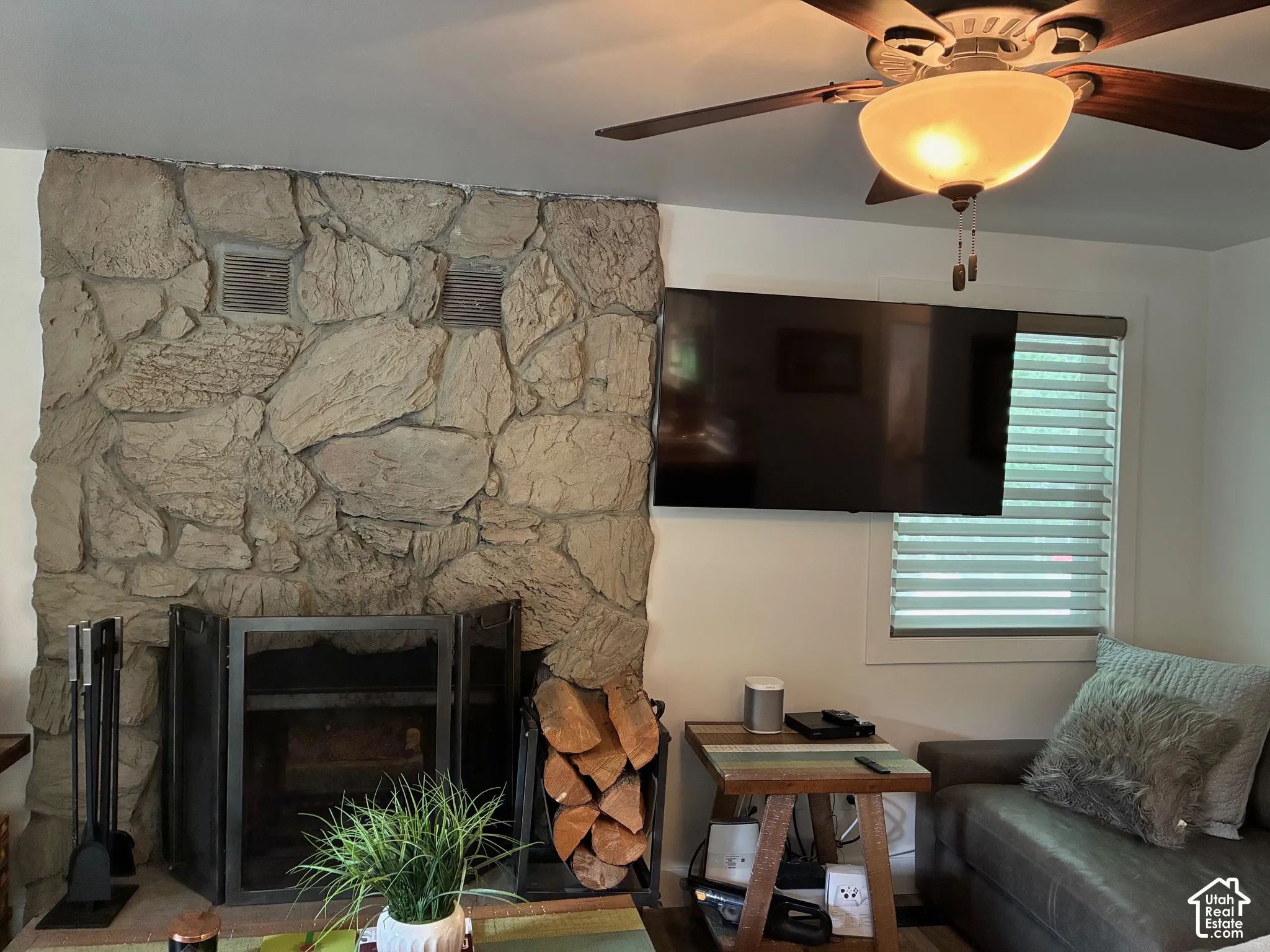 Floor-to-ceiling textured stone fireplace surround with a metal fire screen, ceiling-mounted fan with integrated light fixture, and window with adjustable horizontal blinds