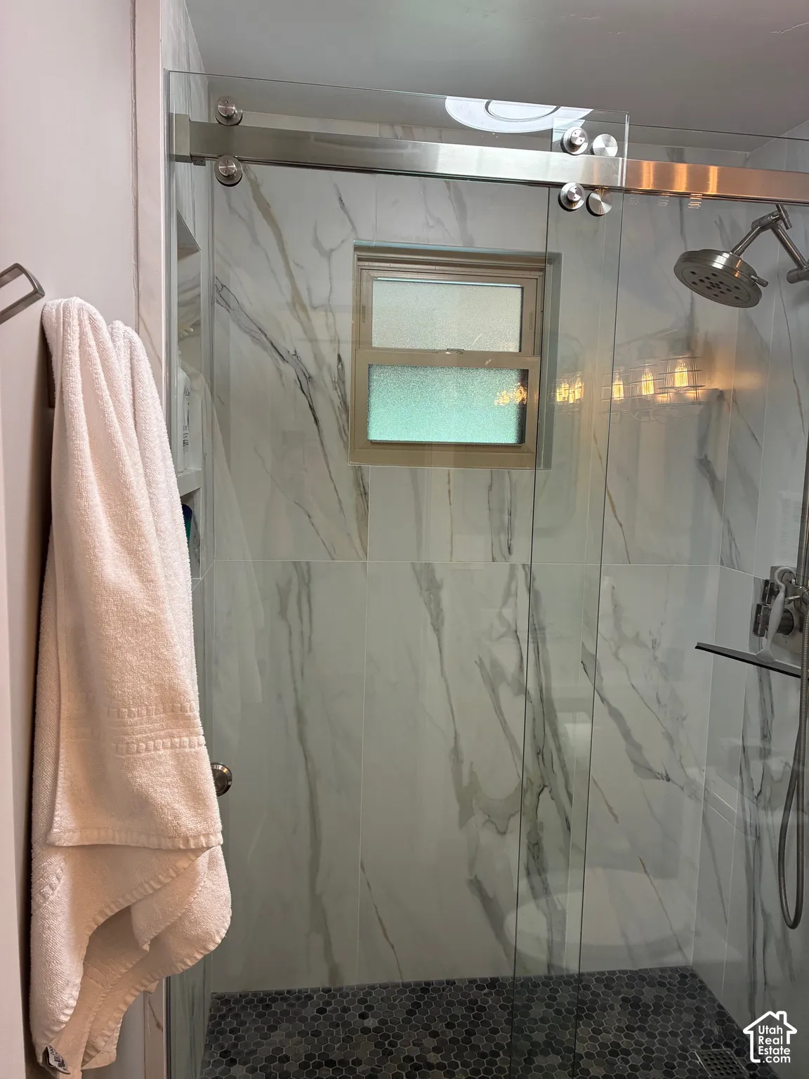Shower enclosure featuring large-format marbled tile walls, a sliding glass door with polished metal hardware, a built-in recessed shelf, and a dark mosaic pebble-tile floor