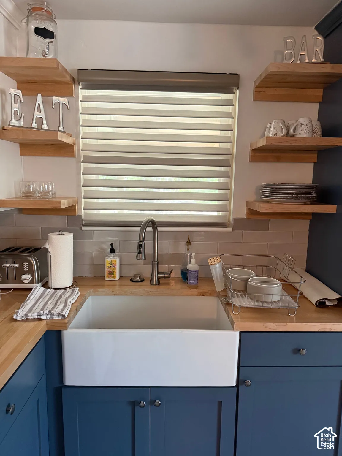 White farmhouse-style apron sink, butcher block countertops, custom blue shaker-style cabinetry, natural wood floating shelves, and subway tile backsplash