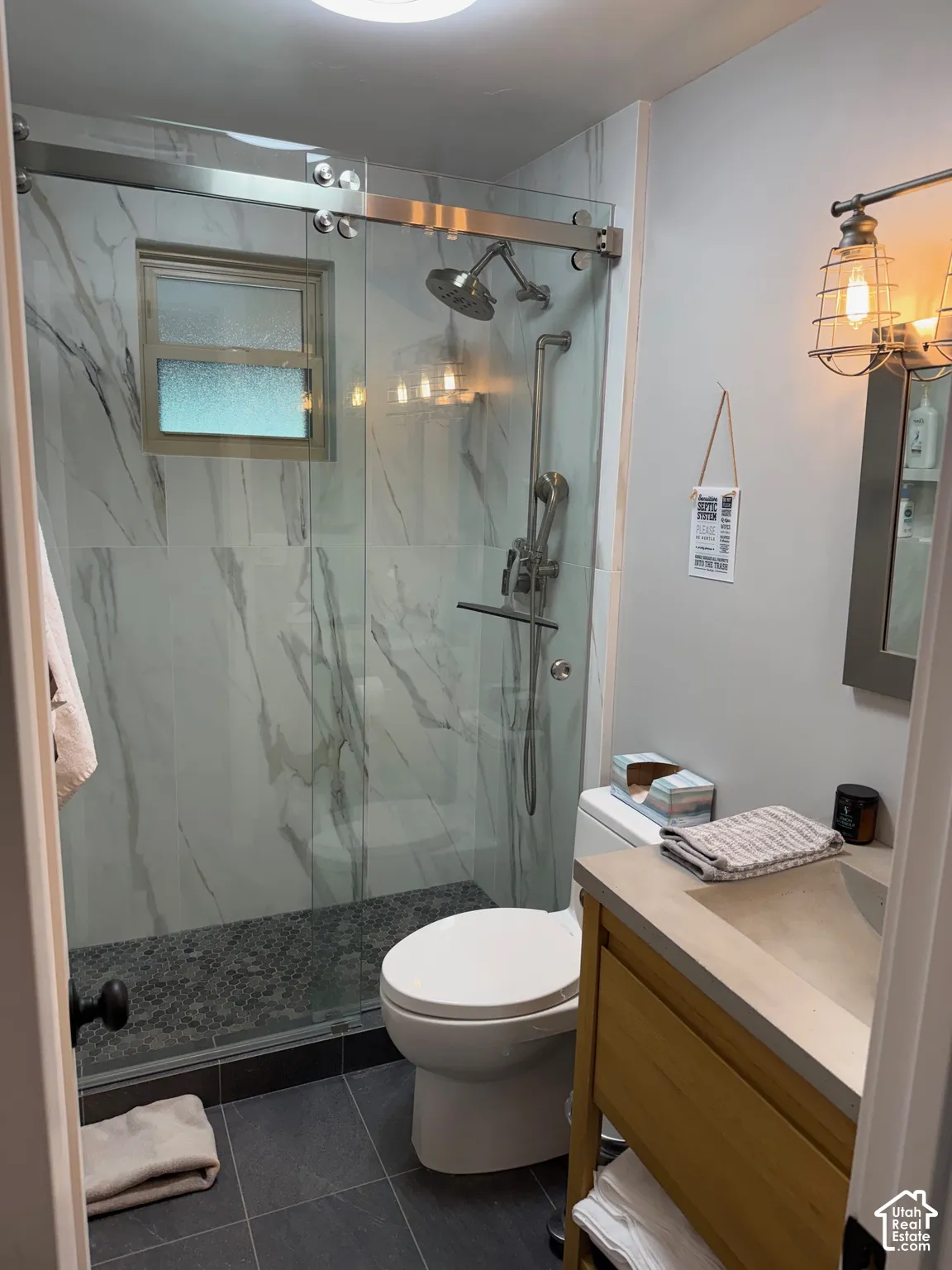 Walk-in shower featuring floor-to-ceiling stone-pattern tile, a sliding glass door, and a dual-head shower system, complemented by a wood-finish vanity with an integrated countertop sink and industrial-style wall lighting