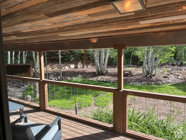Covered deck featuring a natural wood-plank ceiling, wire-mesh railing, and exterior lighting, overlooking a landscaped yard with mature trees
