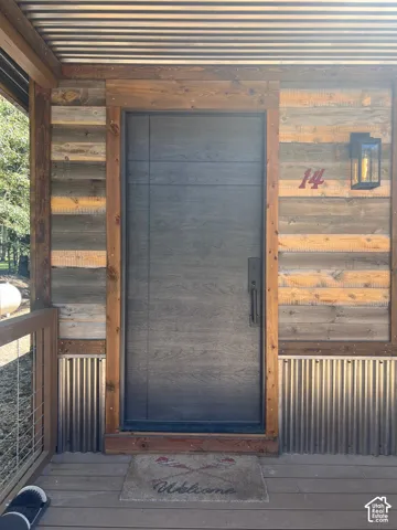 Entryway featuring horizontal wood-plank siding, corrugated metal accents, a dark-finish door with architectural handle hardware, and a wall-mounted exterior sconce