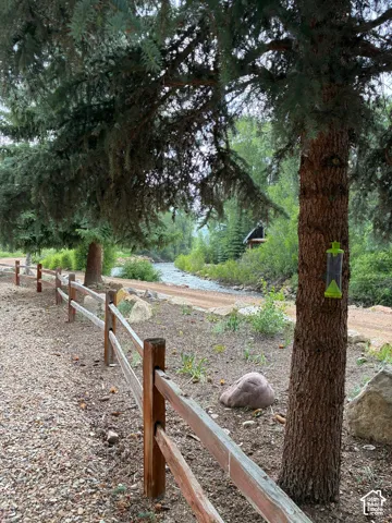 Natural landscape featuring mature evergreen trees, a split-rail wooden fence, and a gravel pathway along a flowing creek