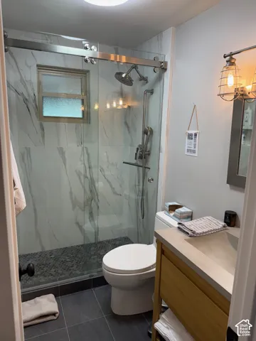 Walk-in shower featuring floor-to-ceiling stone-pattern tile, a sliding glass door, and a dual-head shower system, complemented by a wood-finish vanity with an integrated countertop sink and industrial-style wall lighting