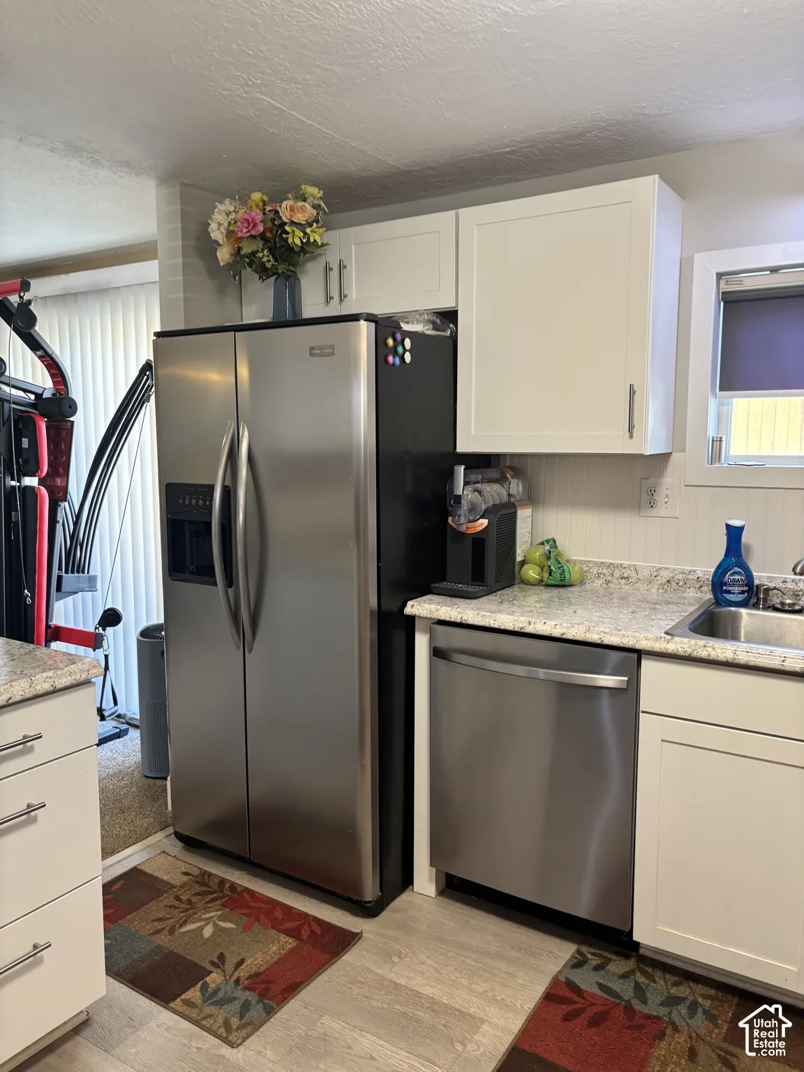 Kitchen area featuring white shaker-style cabinetry, stainless steel side-by-side refrigerator, stainless steel dishwasher, stone-finish countertops, and light-tone wood-finish flooring