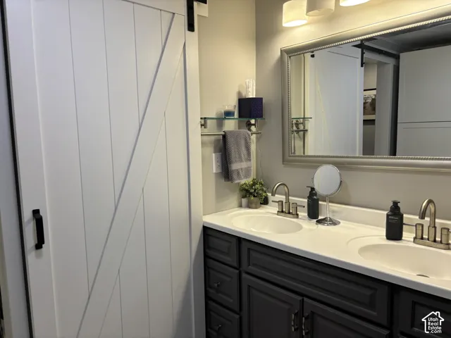 White barn-style sliding door, dual-sink vanity with dark cabinetry, integrated light-colored countertop, framed wall mirror, and metallic finish plumbing fixtures