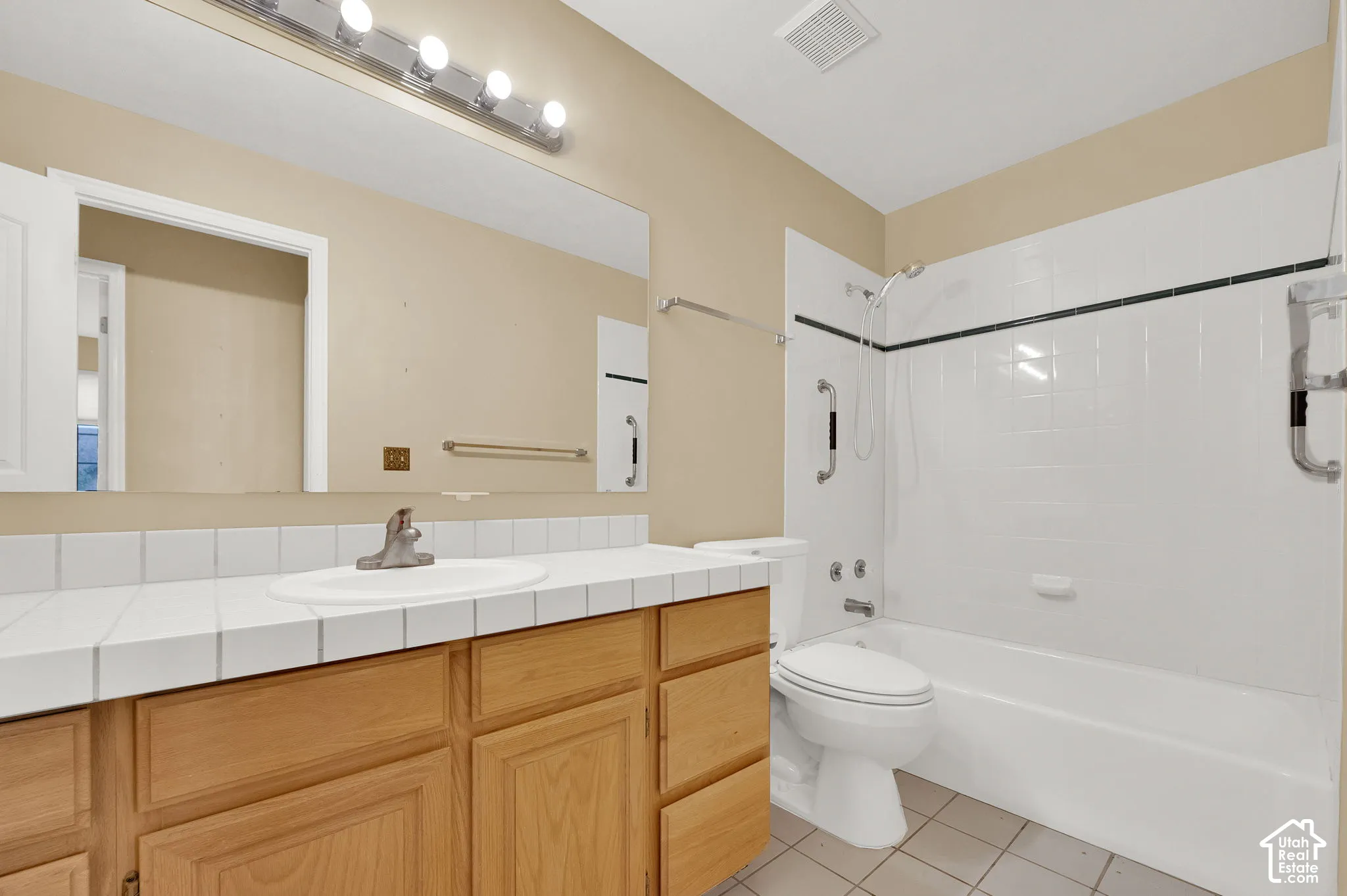 Bathroom featuring a vanity with a white tiled countertop, integrated basin, natural-finish wood cabinetry, a large wall-mounted mirror, and a tub-shower combination with white wall tile