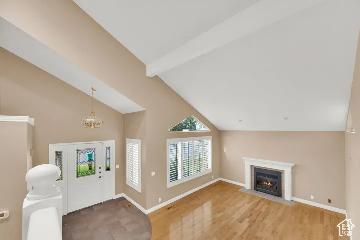 Vaulted ceilings featuring white structural beams, wood-finish flooring, a fireplace with a white mantel, an entryway with tile flooring, and windows with white plantation shutters