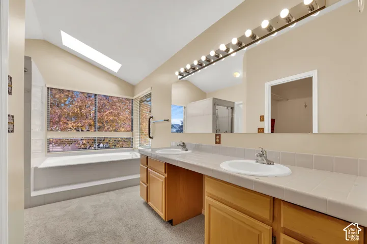 Bathroom featuring a dual-sink vanity with tile countertop, vaulted ceiling with skylight, and a sunken soaking tub