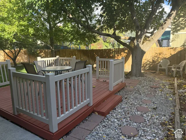 Backyard featuring a raised wooden deck with railings, stone-paver walkway, gravel landscaping, and mature trees enclosed by a wooden fence