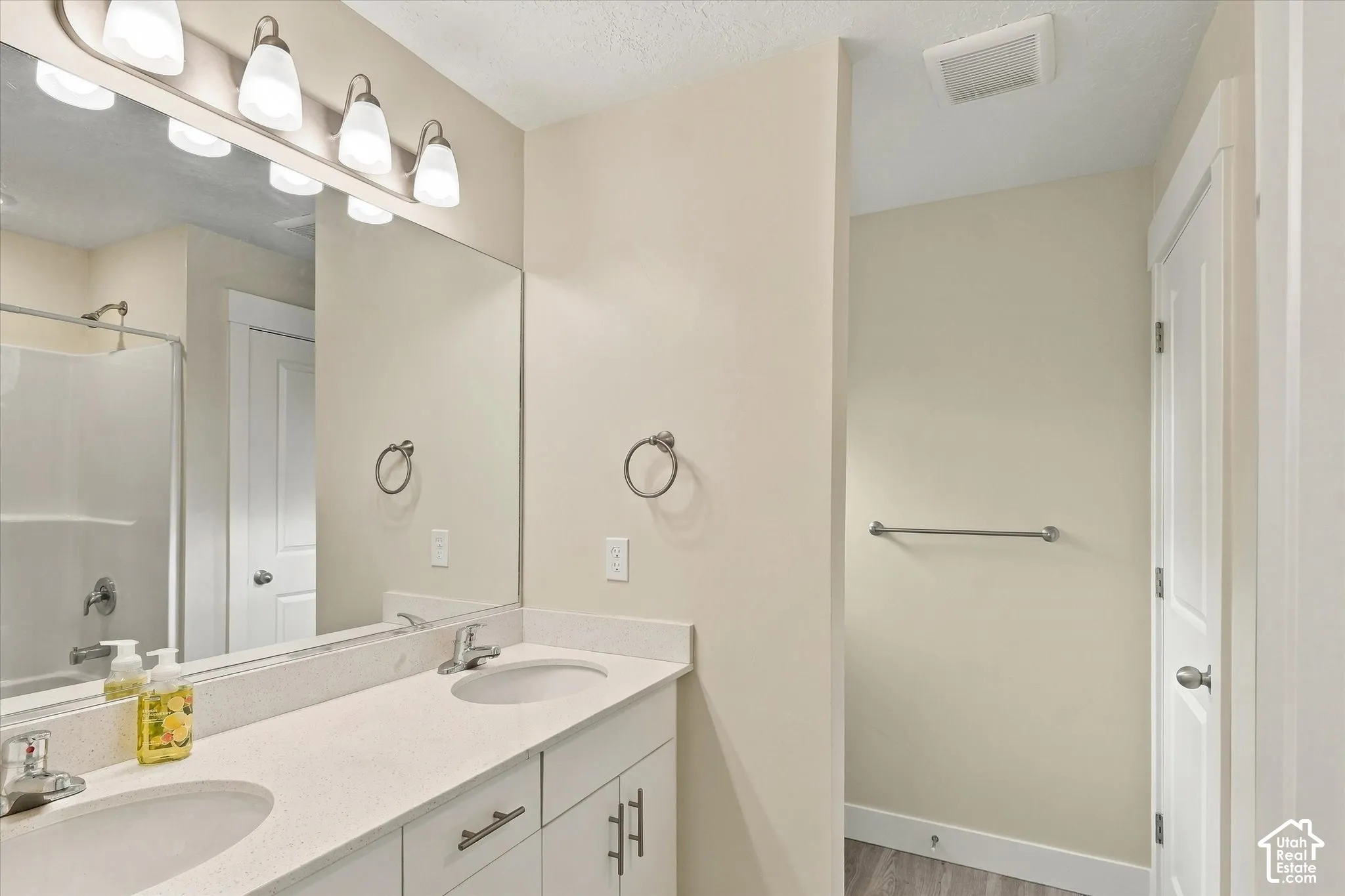 Primary bathroom featuring double vanity with light-toned countertop, integrated dual sinks, polished chrome hardware, and an expansive wall-to-wall mirror with four-bulb overhead lighting