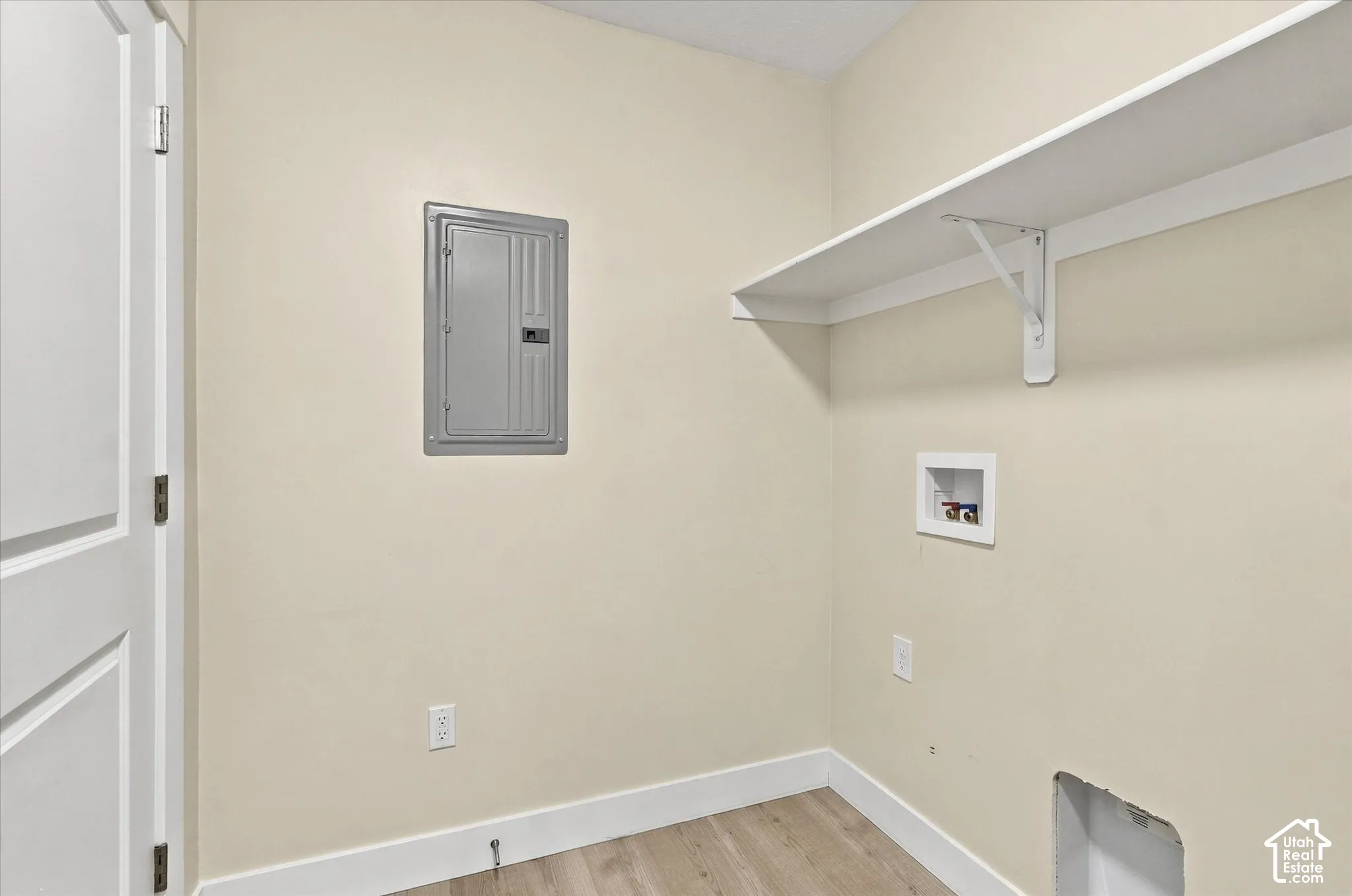 Utility area featuring wood-finish flooring, a wall-mounted electrical panel, a built-in wall shelf with support brackets, and recessed plumbing connections