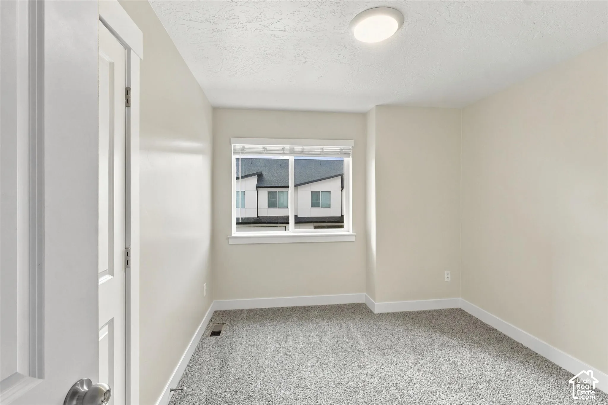 Neutral-toned bedroom featuring light-colored wall paint, plush carpeting, a single-hung window with blinds, and a flush-mount ceiling light fixture