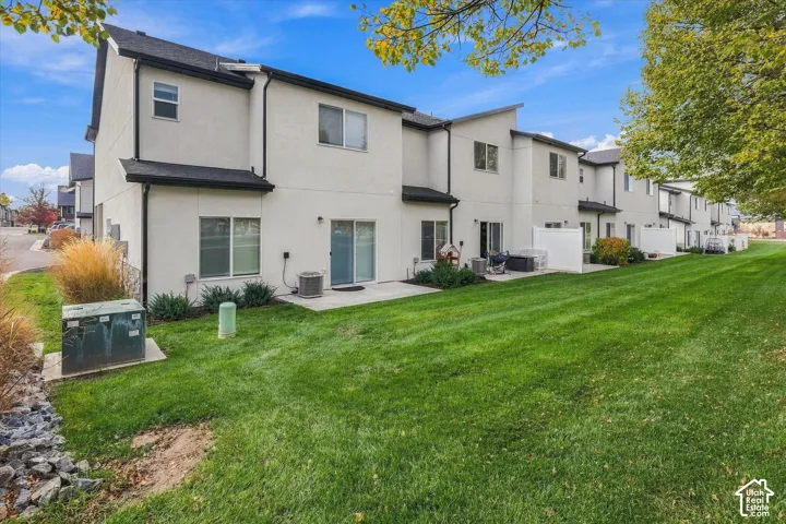 Multi-family exterior featuring light-toned stucco siding, dark metal roof trim, concrete ground-level patios, and expansive shared lawn space
