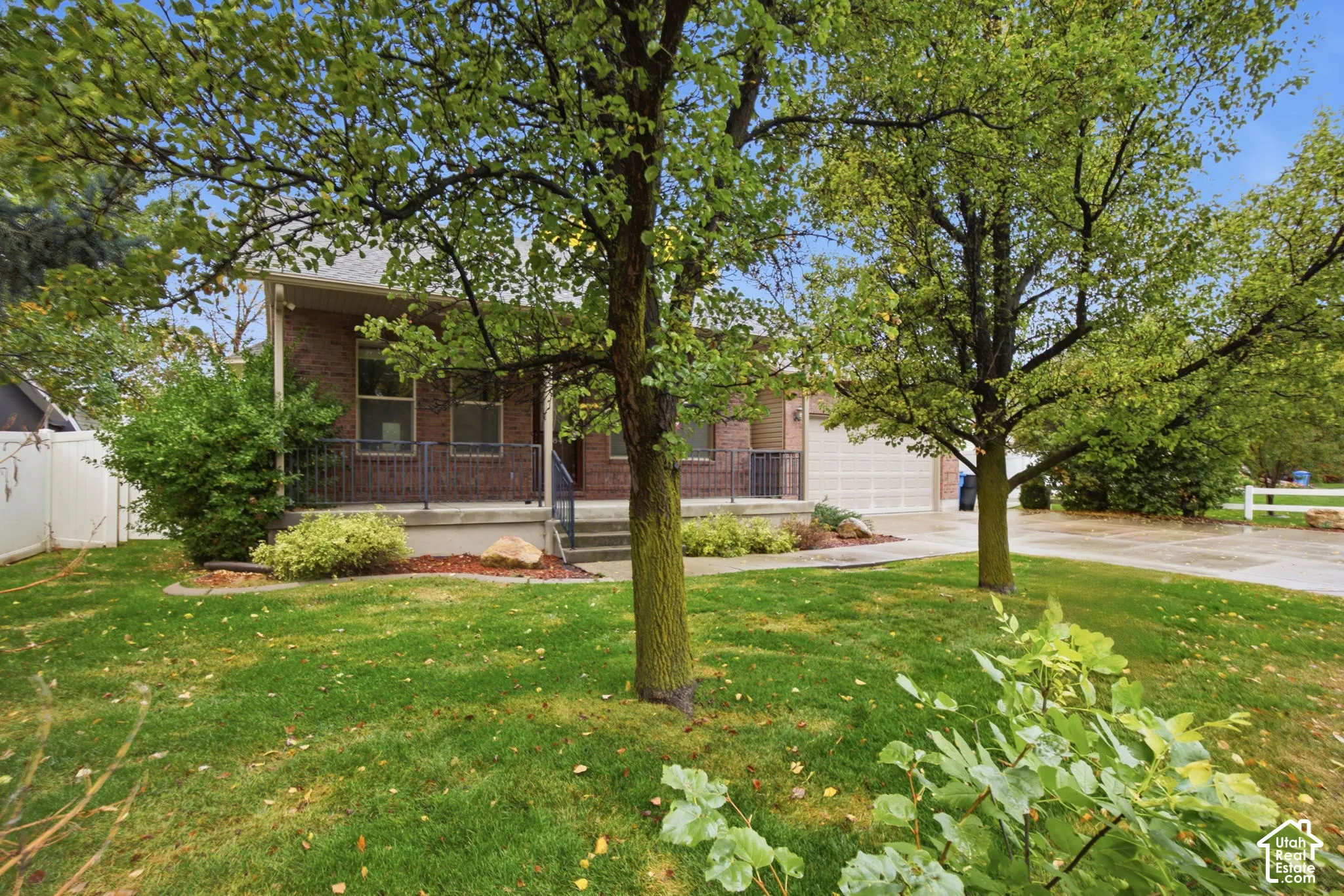 Brick exterior featuring a concrete porch, black metal railings, an attached garage, and mature trees within the front lawn