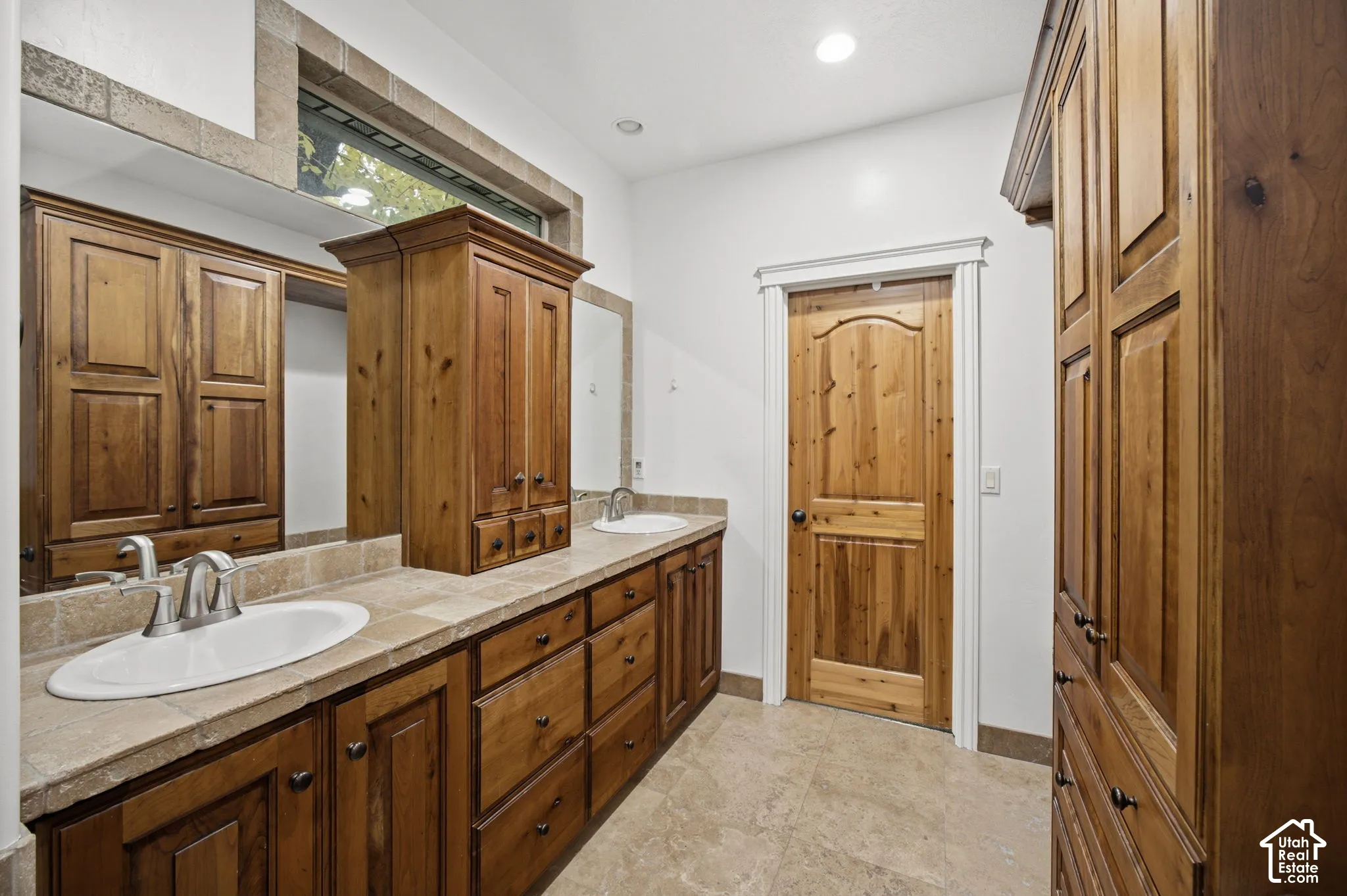 Dual-sink vanity with natural stone tile countertop, custom wood cabinetry, integrated storage tower, and tile flooring