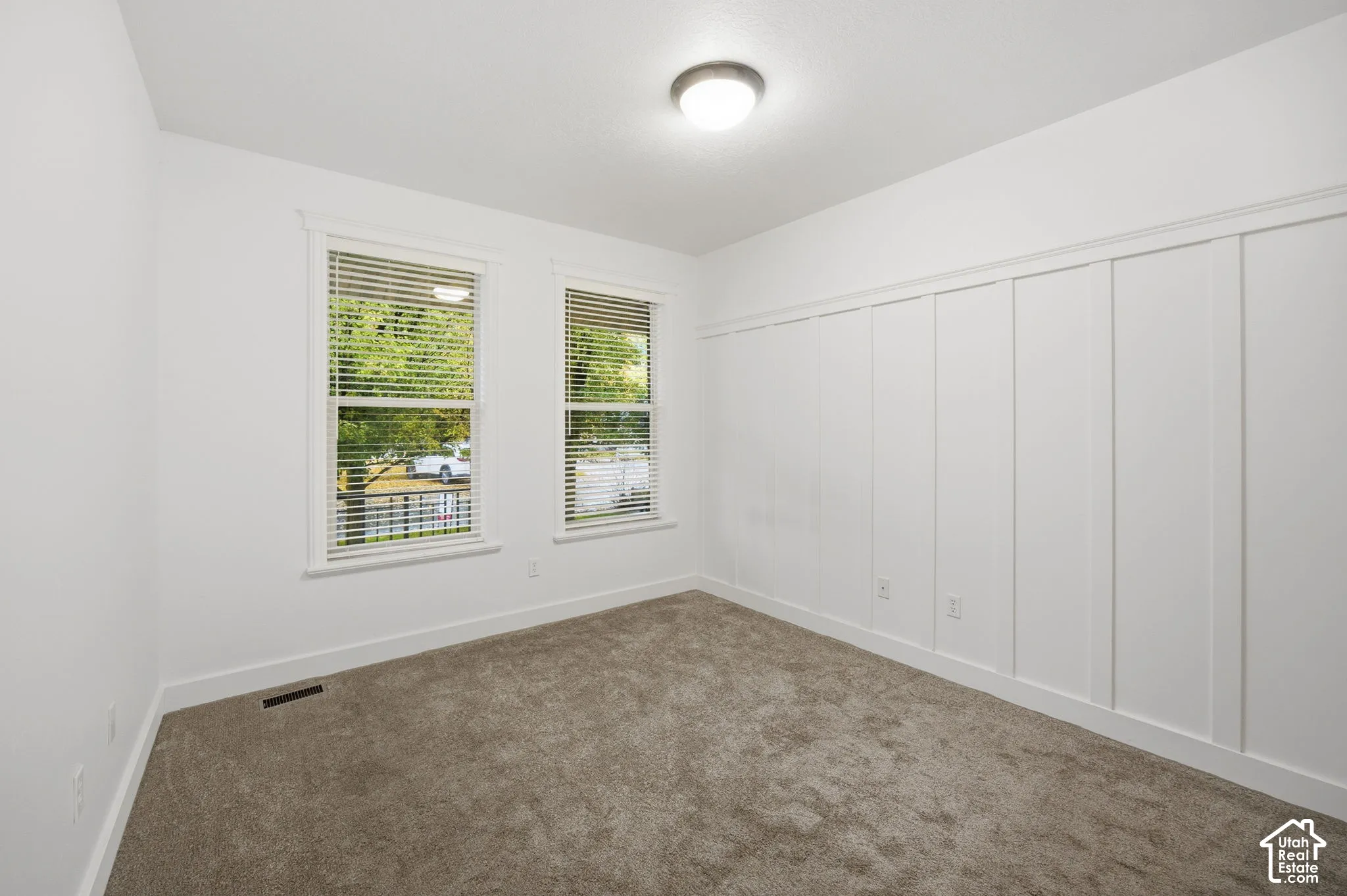 Interior room featuring vertical board-and-batten wall detailing, neutral carpeting, dual windows with installed blinds, and a central flush-mount ceiling light fixture