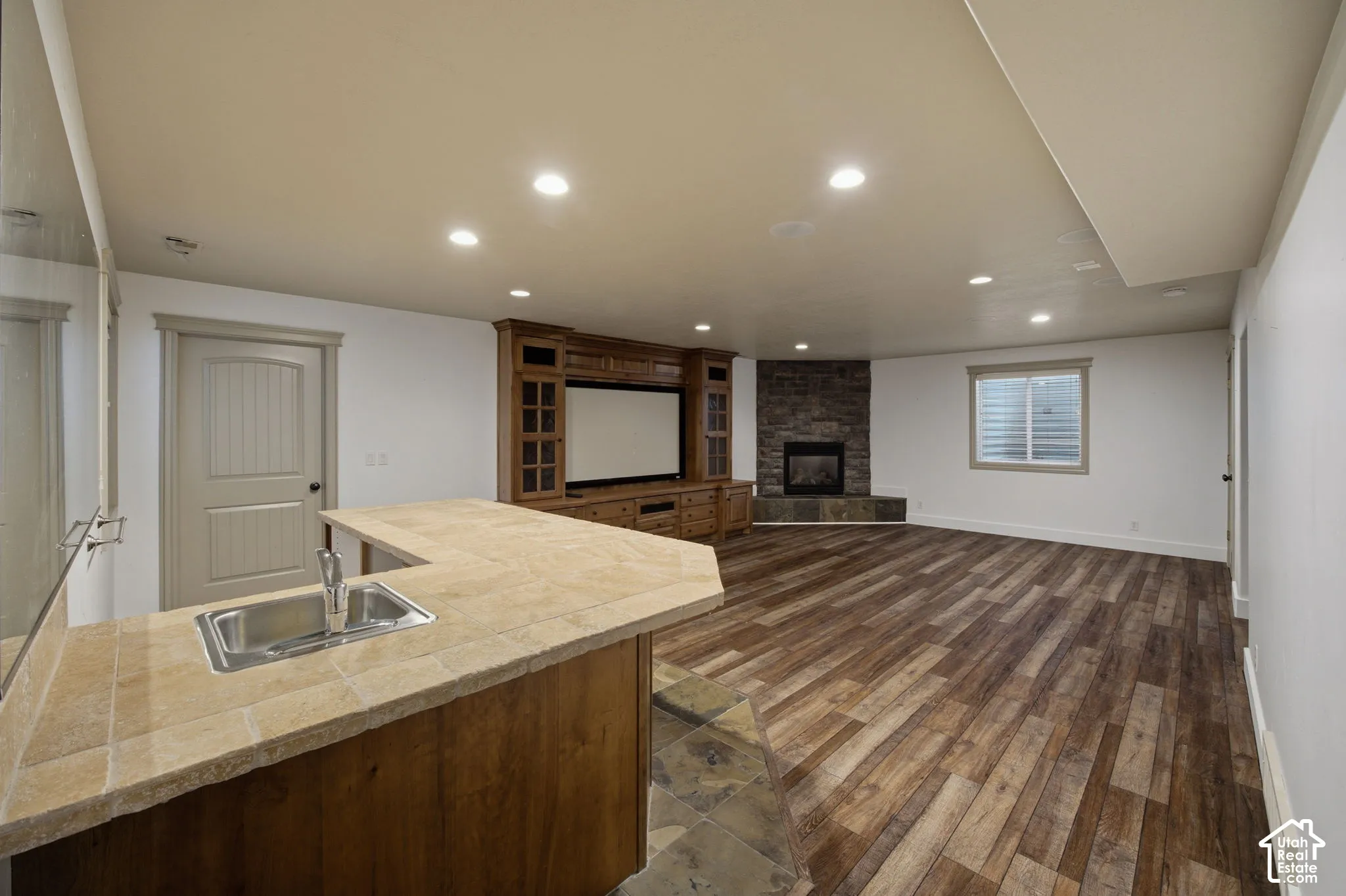 Built-in cabinetry with integrated media center, stone-surround fireplace, tile-topped wet bar with stainless steel sink, wood-finish flooring, and recessed lighting